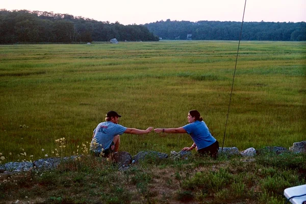 Two people reaching toward each other beside a wide green marsh.